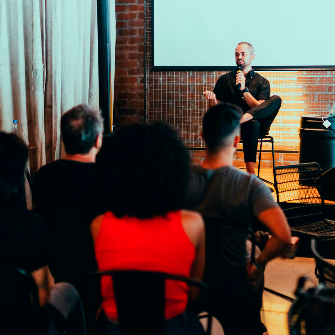 Imagem de pessoa no palco palestrando para alguns espectadores.